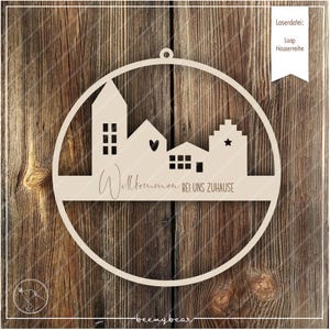 May include: A wooden circle with a cutout design of houses and the text "Willkommen bei uns zuhause". The circle is attached to a string for hanging.