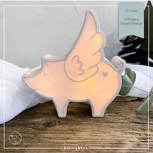 Può includere: Una statuetta a forma di maiale rosa illuminata, con ali d'angelo e bordi bianchi. Il maiale ha un dettaglio a forma di cuore e una coda arricciata. L'immagine include il testo "STL-Datei: Lichterglanz Schweinchenengel" e il logo "beemybear".