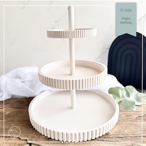 May include: A three-tiered, off-white serving tray with a central support post. Each tier has a circular design with a ribbed edge. The text "STL-Datei: Etagere dreistöckig" is visible in the upper right corner. The tray is on a wooden surface.