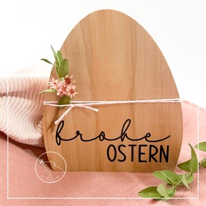 以下が含まれることがあります： 黒い文字で「frohe Ostern」と書かれた木製のイースターエッグ型の看板。看板はピンクの花と白いリボンで飾られています。
