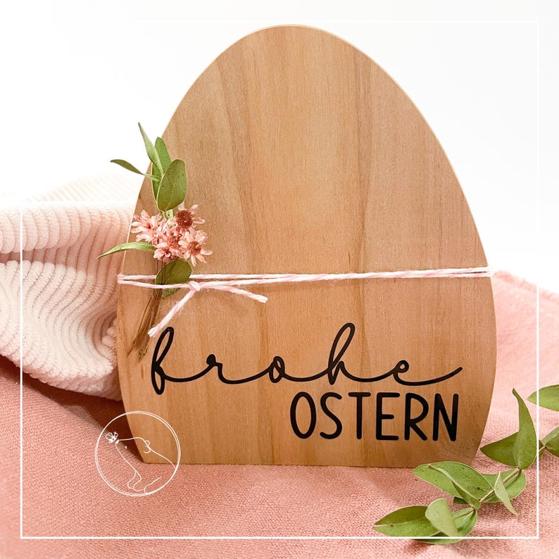 以下が含まれることがあります： 黒い文字で「frohe Ostern」と書かれた木製のイースターエッグ型の看板。看板はピンクの花と白いリボンで飾られています。