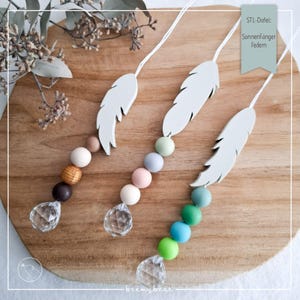 Könnte beinhalten: Drei dekorative Sonnenfänger mit weißen Federanhängern, bunten Perlen und Kristallprismen. Die Perlen sind in verschiedenen Farben gehalten, darunter Weiß, Braun, Rosa, Blau und Grün. Auf einer Holzoberfläche präsentiert.