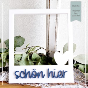 Könnte beinhalten: Ein weißer Bilderrahmen mit einem Herz-Ausschnitt und dem blauen Text "schön hier". Der Rahmen ist mit grünen Blättern und Zweigen dekoriert. Ein türkisfarbenes Etikett oben rechts enthält deutschen Text.