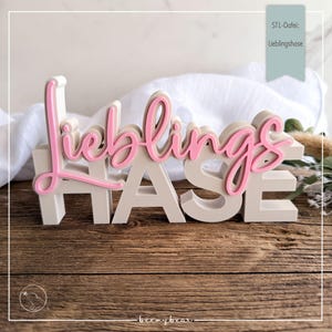Könnte beinhalten: Ein Holzschild mit dem Wort "Lieblingshase" in rosa Schrift über weißen Blockbuchstaben. Das Schild steht auf einer Holzoberfläche mit einem weißen Tuch im Hintergrund. Der Text "STL-Datei: Lieblingshase" befindet sich in einem türkisfarbenen Banner.