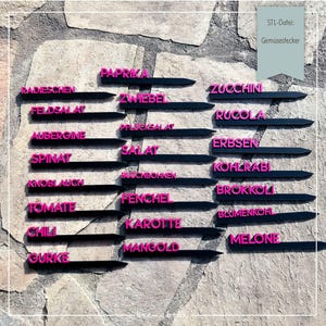 May include: Black garden markers with pink lettering, each labelled with a different vegetable name. The markers are arranged on a stone wall. The text "STL-Datei: Gemüsestecker" is in the upper right corner.