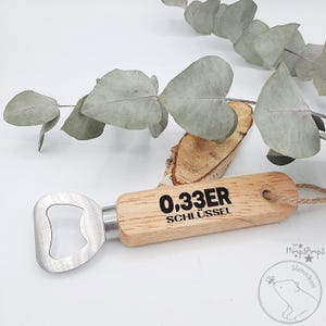 May include: Wooden bottle opener with a metal bottle opener attached. The wooden handle is engraved with the text "0.33ER SCHL&Uuml;SSEL".