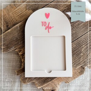 Op de afbeelding: Een witte, boogvormige memo-houder met een rechthoekig venster. De woorden "TO do" staan in roze script, met een roze hart erboven. De houder staat op een houten oppervlak. Tekst in de rechterbovenhoek luidt "STL-Datei: Klebezettelhalter to do".