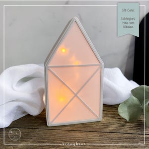 May include: A house-shaped decorative light with a soft pink interior and a white frame. The light is illuminated with small yellow lights, creating a warm glow. The text on the image reads "STL-Datei: Lichterglanz Haus vom Nikolaus."