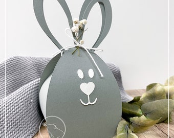 May include: A gray paper bunny shaped box with white bunny ears and a white face. The box is tied with a white string and has a small bouquet of white flowers on top.