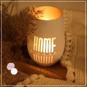 May include: White ceramic candle holder with the words "Home Sweet Home" cut out. The candle is lit and glowing.