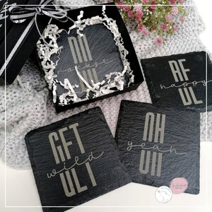 May include: Set of four black slate coasters with white lettering. The coasters feature the phrases "Get  wed  ULT", "Oh yeah ULT", "AF happy ULT", and "NI  rockets  ULT". The coasters are packaged in a black gift box with white tissue paper.