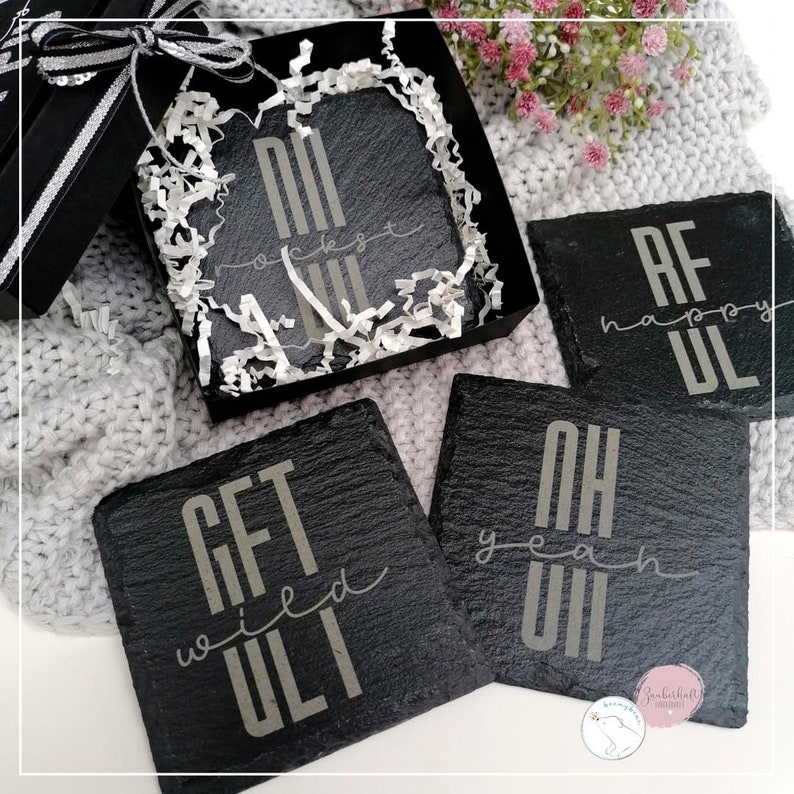 May include: Set of four black slate coasters with white lettering. The coasters feature the phrases "Get  wed  ULT", "Oh yeah ULT", "AF happy ULT", and "NI  rockets  ULT". The coasters are packaged in a black gift box with white tissue paper.