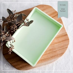 May include: A rectangular, light green tray sits on a wooden cutting board. The tray is empty and has a slightly raised edge. Dried foliage with brown leaves and small buds is placed on the left side. The text "STL-Datei: Schale Rechteck" is in the upper right corner.