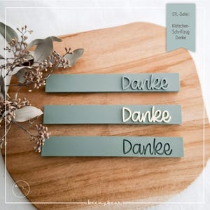 Könnte beinhalten: Drei rechteckige, salbeigrüne Tafeln mit dem Wort "Danke" in einer dekorativen Schriftart. Die Tafeln sind auf einem Holzschneidebrett angeordnet, mit getrockneten floralen Akzenten. Der Text "STL-Datei: Klötzchen-Schriftzug Danke" befindet sich oben rechts.