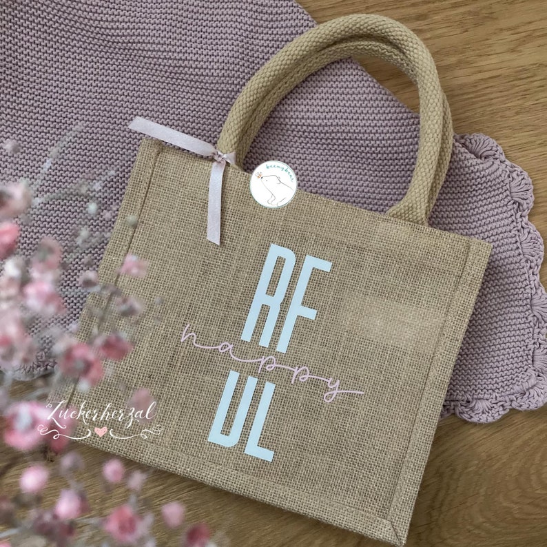 May include: A brown burlap tote bag with white fabric handles. The bag has the letters "RF" in large white letters and the word "happy" written in pink cursive script. The letters "UL" are also in large white letters. The bag is sitting on a pink knitted blanket.