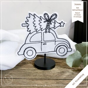 Op de afbeelding: Een witte vilten auto met zwarte geborduurde contouren, met een kerstboom vastgebonden met een strik. De auto is gemonteerd op een zwarte standaard. De afbeelding bevat Duitse tekst.