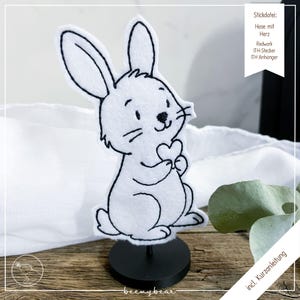 May include: A white felt bunny figurine holding a heart, with black outline details. The bunny is mounted on a black stand. The image includes text in German: "Stickdatei: Hase mit Herz, Redwork, ITH-Stecker, ITH Anhänger."