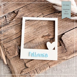 May include: A white rectangular photo frame with a square opening, featuring a small white heart shape in the bottom right corner and the light blue text "Fellnase" embossed on the bottom border. The frame has a clean, flat design.