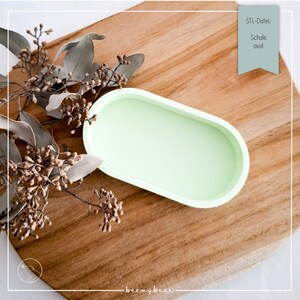 May include: A pale green oval tray on a wooden board. The tray has a smooth finish and a raised edge. Dried flowers are on the left. Text on the right says "STL-Datei: Schale oval".