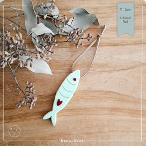 Puede incluir: Colgante en forma de pez azul claro con un corazón y un ojo rojos, colgado de un cordón beige. El colgante tiene un diseño minimalista. El fondo incluye follaje seco y el texto "STL-Datei: Anhänger Fisch".
