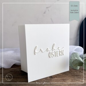 Könnte beinhalten: Eine weiße Karte mit den Worten "Frohe Ostern" auf der Vorderseite geprägt. Die Karte ist leicht angewinkelt und zeigt ihr gefaltetes Design. Der Hintergrund umfasst eine Holzoberfläche und einen verschwommenen Hintergrund.