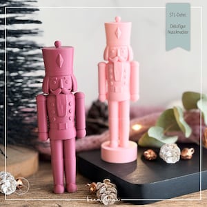 May include: Two nutcracker figurines in matte finishes, one in a deep mauve and one in a light pastel pink. Each figure features a tall hat, a uniform-style jacket with buttons, and a mustache. A label reads STL-Datei: Dekofigur Nussknacker.