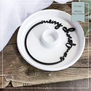 May include: A white ceramic plate with a black circular design and the words "merry christmas" in a cursive font. The plate has a small, raised centre and sits on a wooden surface. The text "STL-Datei: Tellerkranz Merry Christmas" is also visible.