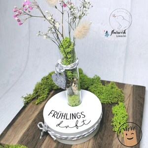 以下が含まれることがあります： 黒い文字で「FR&Uuml;HLINGS duft」と書かれた白い木製の台座。苔と小さな白い花で満たされたガラス製の試験管が台座に差し込まれています。台座は緑の苔で囲まれています。
