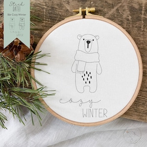 May include: A white embroidery hoop with a black and white illustration of a bear wearing a scarf. The text "cozy winter" is embroidered below the bear.
