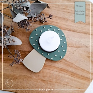 May include: A green mushroom-shaped candle holder with a white LED candle. The holder has small, light-colored dots around the edge and a beige stem. The text "STL-Datei: Teelichthalter Fliegenpilz" is visible on a teal tag.