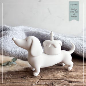 Peut inclure: Une figurine décorative blanche mate représentant un chien teckel avec un petit gâteau surmonté d'une bougie sur le dos. Le texte sur l'étiquette indique : STL-Datei: Dekofigur Dackel Franz mit Törtchen. Le chien a une finition lisse.