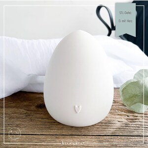 Peut inclure: Un grand objet décoratif blanc en forme d'œuf avec une petite indentation en forme de cœur. L'objet est posé sur une surface en bois. Le texte sur une bannière indique "STL-Datei: Ei mit Herz".