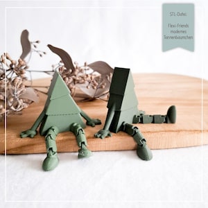 Op de afbeelding: Twee groene, beweegbare boomfiguren met armen en benen zitten op een houten oppervlak. De bomen hebben een geometrisch ontwerp en bevinden zich in een ontspannen pose. De afbeelding bevat de tekst "STL-Datei: Flexi-Friends modernes Tannenbäumchen."