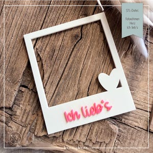 Può includere: Cornice bianca in stile Polaroid con un cuore ritagliato e la scritta rosa "Ich lieb's". La cornice è posizionata su una superficie di legno. Cornice decorativa per foto.