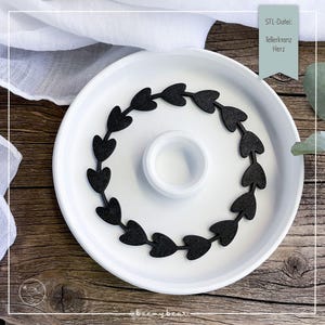 Könnte beinhalten: Ein weißer Keramikteller mit einem schwarzen herzförmigen Girlande am Rand. Der Teller hat eine zentrale, erhöhte kreisförmige Fläche. Der Text "STL-Datei: Tellerkranz Herz" ist ebenfalls vorhanden.