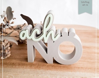 May include: A gray concrete sculpture of the word "ach nö" with a light green accent. The letters are in a playful font, with the "ach" in cursive and the "nö" in block letters. The sculpture is on a wooden surface.