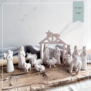 May include: A matte white nativity set featuring figurines of an angel, figures of Mary and Joseph, the baby Jesus in a manger, three wise men, a shepherd, a cow, a donkey, and two sheep, all positioned under a simple stable structure with a star.