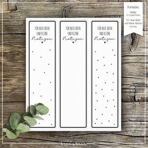 May include: Three white rectangular bookmarks with black borders, each featuring the text "FÜR NEUE IDEEN UND KLEINE Notizen" and small black heart and dot designs. The image includes text in German and a sprig of green leaves.