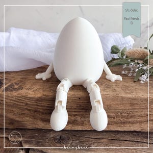 Peut inclure: Figurine blanche en forme d'œuf avec des jambes et des bras articulés, posée sur une surface en bois. La figurine a un design fantaisiste, avec le texte "STL-Datei: Flexi-Friends Ei" sur une bannière turquoise. La marque "beemybear" est en bas.
