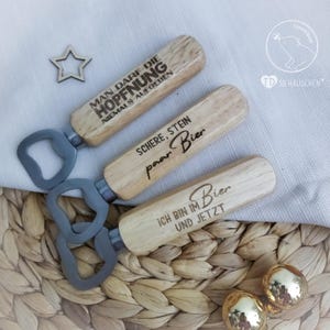 May include: Three wooden bottle openers with engraved sayings. The sayings are "Man darf die Hoffnung niemals aufgeben", "Schere, Stein, paar Bier", and "Ich bin im Bier und jetzt".