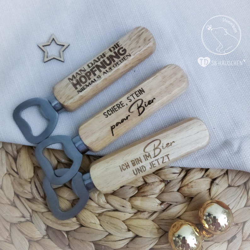 May include: Three wooden bottle openers with engraved sayings. The sayings are "Man darf die Hoffnung niemals aufgeben", "Schere, Stein, paar Bier", and "Ich bin im Bier und jetzt".