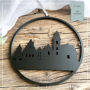 Könnte beinhalten: Ein schwarzer Metallschmuck mit einer Silhouette eines Dorfes in einem kreisförmigen Rahmen. Das Design umfasst Häuser, eine Kirche und Bäume. Der Schmuck hängt an einem weißen Band. Der Text "STL-Datei: Loop Winterdorf" ist sichtbar.