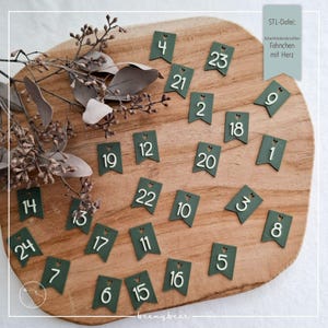May include: A wooden cutting board displays 24 numbered green tags for an advent calendar. Each tag has a white number and a heart-shaped cutout. The text "STL-Datei: Adventskalenderzahlen Fähnchen mit Herz" is in the upper right corner.