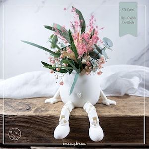May include: A white, egg-shaped vase with articulated legs and arms, holding a bouquet of pink and green flowers. The vase has a heart detail and the text "STL-Datei: Flexi-Friends Eierschale". The vase is sitting on a wooden surface.