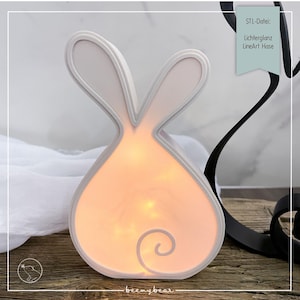 May include: A white bunny-shaped night light with a soft, glowing interior. The light has a curved design and a swirl detail at the bottom. The text "STL-Datei: Lichterglanz LineArt Hase" is on a teal tag. The base is on a wooden surface.