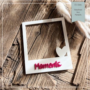 Op de afbeelding: Een witte fotolijst met een hartuitsparing en het woord "Moments" in roze script. De lijst staat op een rustiek houten oppervlak, met de tekst "STL-Datei: Fotorahmen Herz Moments" rechtsboven.