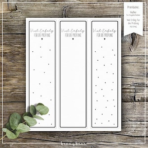 May include: Three white rectangular bookmarks with black borders, each featuring the text "Viel Erfolg FÜR DIE PRÜFUNG" and small black hearts. The bookmarks are decorated with small black dots and are on a wooden surface.