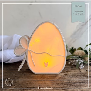 Peut inclure: Lampe en forme d'œuf, émettant une lumière orange douce. Elle est ornée d'un nœud décoratif et d'un petit cœur. Le texte sur l'image indique "STL-Datei: Lichterglanz Ei mit Schleife".