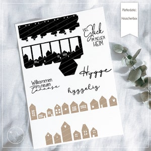 Könnte beinhalten: Ein druckbares Designblatt mit schwarzen und braunen Silhouetten von Häusern, Sternen und Text. Der Text beinhaltet "Viel Glück im neuen Heim", "Willkommen im neuen Zuhause", "Hygge" und "hyggelig".