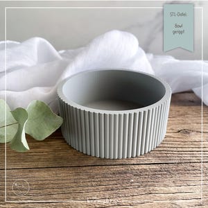 Op de afbeelding: Een grijze, geribbelde betonnen kom. De kom heeft een cilindrische vorm met verticale ribbels aan de buitenkant. De tekst "STL-Datei: Bowl gerippt" staat op een blauwgroen label. De kom staat op een houten oppervlak met witte stof en groene bladeren op de achtergrond.